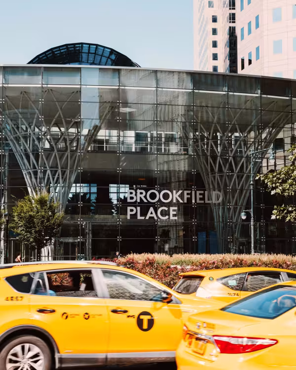Street-level view of Brookfield Place New York, a BGRE operated property in Lower Manhattan, featuring a glass entrance pavilion and vibrant urban streetscape with passing yellow taxis.